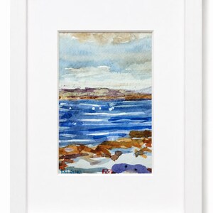 Puede incluir: Una acuarela de un paisaje costero con un mar azul profundo, una costa rocosa de color marrón y un cielo pálido, encerrada en un marco rectangular blanco sencillo con un borde de paspartú blanco. La obra incluye una firma en la esquina inferior izquierda.