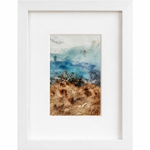 Puede incluir: Una pintura en acuarela enmarcada que presenta un paisaje abstracto con un primer plano marrón texturizado y un cielo vibrante en azul y blanco. La obra está contenida en un marco rectangular blanco con un borde de paspartú blanco.