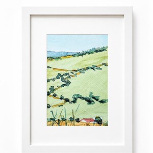 Puede incluir: Una pintura de acuarela enmarcada que representa un paisaje verde ondulado con setos de color verde oscuro, un cielo azul claro y un toque de colinas azules distantes. La obra está encerrada en un marco rectangular blanco simple con paspartú.