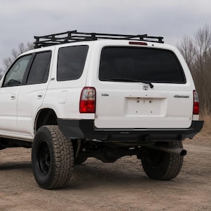 Pode incluir: Um SUV Toyota 4Runner branco com bagageiro preto, vidros traseiros fumê e pneus off-road pretos grandes. O veículo está equipado com um para-choque traseiro preto e um cano de descarga visível em um caminho de terra.