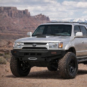 Pode incluir: Um SUV Toyota 4Runner prateado com para-choque dianteiro preto de reposição com suporte para guincho, rodas off-road pretas com pneus de cravos, entrada de ar no capô e rack de teto. O veículo está posicionado sobre um solo de terra.