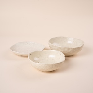 May include: A set of three off-white ceramic dishes featuring a speckled finish. The collection includes one shallow plate and two deeper bowls, all characterised by organic, slightly irregular edges and a smooth, matte-textured surface appearance.