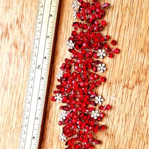 May include: A decorative hair accessory featuring a cascade of red and clear crystals, accented with small white floral details. The piece is set on a gold-toned wire, and is approximately 20 cm long.
