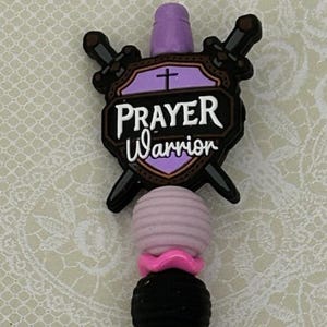Puede incluir: Un bolígrafo morado con las palabras "Prayer Warrior" en la parte superior. El bolígrafo tiene una parte superior decorativa con una cruz y un diseño negro y marrón. El bolígrafo tiene una banda plateada y una punta plateada.
