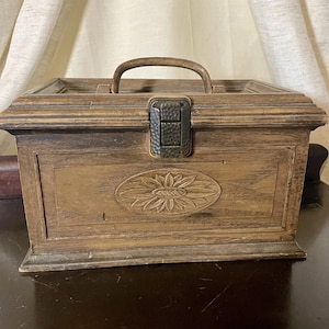 Peut inclure: Une boîte en bois rectangulaire avec une poignée et un fermoir en métal. La boîte présente un motif floral sculpté sur le devant. Le bois a une teinte marron chaude et le fermoir en métal a une finition texturée. La boîte est probablement utilisée pour le stockage.