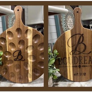 May include: A round wooden serving board with a handle, featuring a carved design of oval indentations. The board is engraved with the letter "B" and the name "Boudreaux" with "est 2017". The wood has a natural grain and a warm brown color.