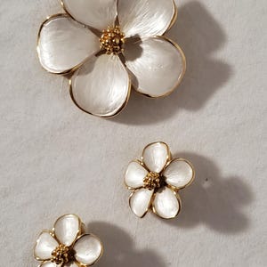 May include: Three white flower-shaped brooches with gold-colored trim and centers. The largest brooch has five petals, while the smaller ones have five petals. The brooches are likely made of metal and enamel.