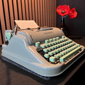 May include: A vintage Hermes 3000 typewriter in a light seafoam green finish with matching mint green keys. The machine features a metal body, a carriage return lever, and a sheet of paper inserted into the platen. The keys display black letters.