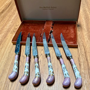 May include: Set of six vintage Sheffield cutlery knives with floral patterned handles in a presentation box. The handles are white with purple and gold floral designs. The box is brown with a red velvet interior.