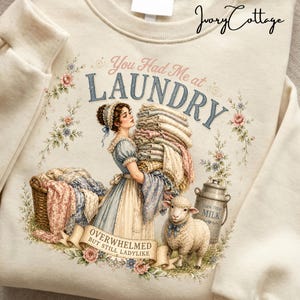 May include: Cream-colored sweatshirt with a vintage-style laundry-themed graphic. The design features a woman carrying laundry, a sheep, and text that reads "You Had Me at Laundry" and "Overwhelmed But Still Ladylike."