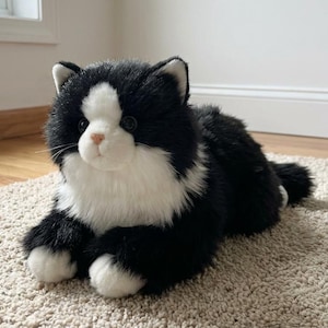 May include: A plush, black and white cat toy with a soft, fluffy texture. The toy cat is lying down on a beige carpet, with its paws and face in white and the rest of its body in black.