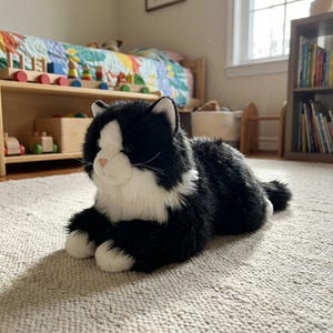 May include: A plush black and white cat toy with a white face, chest, and paws. The cat is lying on a light-colored rug in a child's room. The room has a wooden bookshelf and a bed with a colorful quilt.