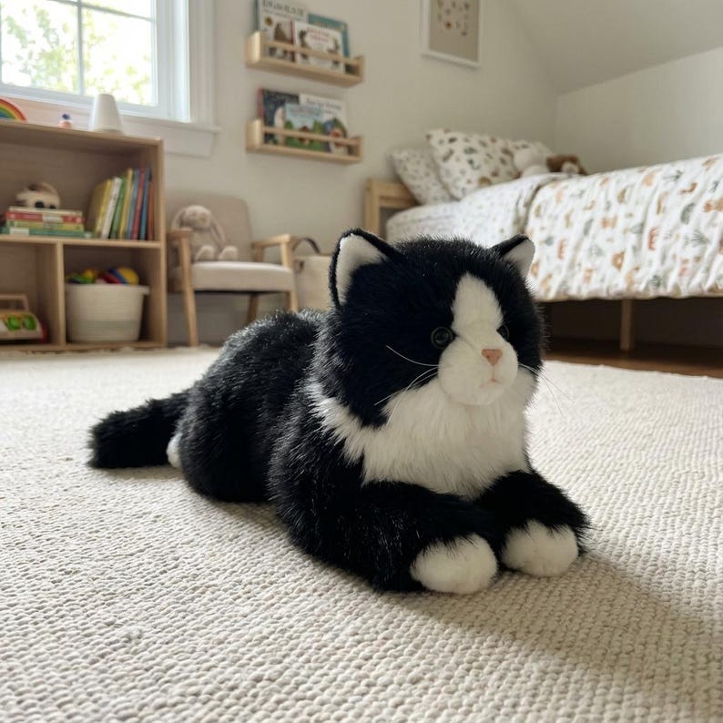 May include: A plush black and white cat toy with a pink nose and white paws. The stuffed animal is lying on a cream-colored rug in a child's bedroom. The cat has a realistic design.