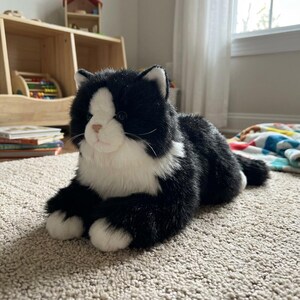 May include: A plush black and white cat toy with a realistic design. The stuffed animal has a white face, chest, and paws, with black fur on the rest of its body. The cat is lying down on a beige carpet.