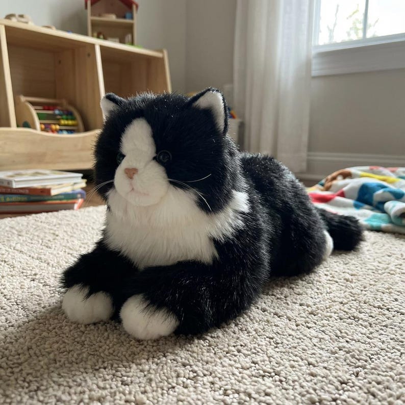 May include: A plush black and white cat toy with a realistic design. The stuffed animal has a white face, chest, and paws, with black fur on the rest of its body. The cat is lying down on a beige carpet.