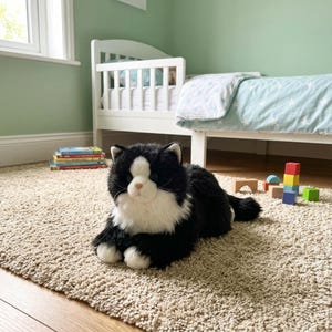 May include: A plush black and white cat toy with a pink nose and white paws is lying on a beige carpet. A white crib, books, and colorful wooden blocks are in the background, suggesting a child's room.