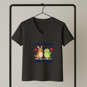 Può includere: T-shirt nera con scollo a V con un canguro e un uccello dei cartoni animati che tengono fiori rossi. Il testo "TWO NATIONS ONE SPIRIT" è stampato in blu. La maglietta è appesa a un attaccapanni nero.
