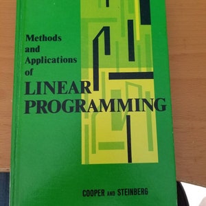 May include: A hardcover book titled "Methods and Applications of Linear Programming" by Cooper and Steinberg. The book has a green cover with a geometric design in shades of green, yellow, and black. The title is in large, bold letters.