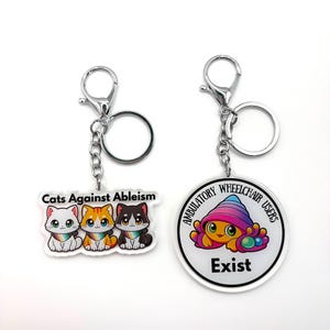May include: Two acrylic keychains with silver-tone metal clasps. The left rectangular keychain features three cartoon cats wearing rainbow collars and the text Cats Against Ableism. The right circular keychain shows a colourful hermit crab and the text Ambulatory Wheelchair Users Exist.