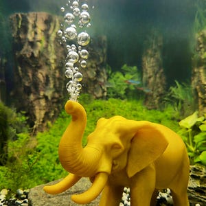 May include: A yellow elephant figurine in an aquarium, with its trunk raised, emitting a stream of bubbles. The background features aquatic plants and a rocky substrate, creating an underwater scene.