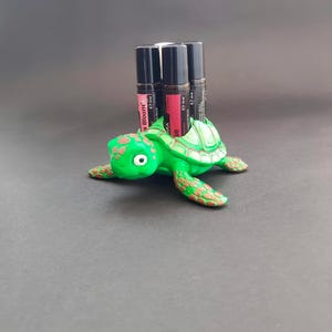 May include: A green turtle figurine with a textured shell and brown accents, holding three essential oil roller bottles. The bottles have black caps and labels with the text "Bloom" and "Ease".