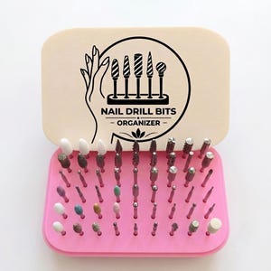 May include: A pink rectangular nail drill bit organizer with a beige lid featuring a black graphic of a hand and five drill bits with the text NAIL DRILL BITS ORGANIZER. The base contains numerous holes holding various metal and ceramic drill bits.