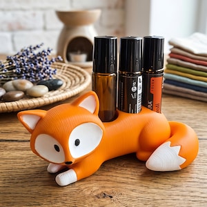 Op de afbeelding: Een oranje en witte vossenvormige houder met drie cilindrische openingen op de rug, die drie donker amberkleurige glazen etherische olie rollerflesjes met zwarte doppen bevat. De vossenfiguur ligt in een liggende positie met een witte staartpunt.