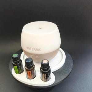 May include: A white and beige dōTERRA essential oil diffuser with a rounded top and two control buttons, accompanied by three 15 ml amber glass bottles labelled Tea Tree, Frankincense, and Cedarwood, all arranged on a circular display stand.