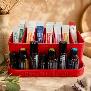 Könnte beinhalten: Ein roter Kunststoff-Organizer mit der Prägung ESSENTIAL OIL auf der Vorderseite. Der Organizer verfügt über mehrere Fächer, in denen verschiedene dōTERRA ätherische Öl-Roll-on-Flaschen und mehrere rechteckige Nahrungsergänzungsmittel-Tüten stehen.