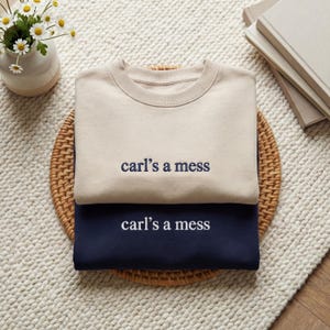 May include: Two crewneck sweatshirts, one beige and one navy blue, are stacked on a woven mat. Each sweatshirt has the embroidered text "carl's a mess" in a contrasting color. The sweatshirts are on a white rug.