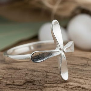 May include: A silver ring featuring a four-petal flower design. The ring is displayed on a piece of weathered wood, with blurred greenery and white stones in the background. The ring's petals have a smooth, shiny finish.