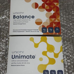 Op de afbeelding: Twee dozen voedingssupplementen van Unicity. De bovenste doos is voorzien van het opschrift "Balance" met "Natuurlijke sinaasappelsmaak" en de onderste doos is voorzien van het opschrift "Unimate" met "Natuurlijke citroensmaak". Beide dozen zijn wit met kleurrijke geometrische ontwerpen.