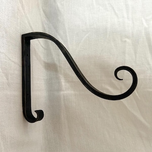Pode incluir: Um suporte de parede preto em ferro forjado para plantas. O suporte apresenta um braço curvo com uma extremidade decorativa em espiral e um gancho na parte inferior. O design é simples e elegante, adequado para exibir plantas suspensas ou outros objetos decorativos.