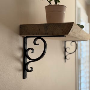 Puede incluir: Estantería rústica de madera con borde natural, soportada por soportes decorativos de metal negro. Una pequeña planta en maceta se encuentra sobre la estantería. La estantería está montada en una pared beige. La estantería mide unos 90 cm.