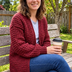 Könnte beinhalten: Eine tiefrote, offene Strickjacke mit strukturiertem Waffelmuster, langen Ärmeln mit gerippten Bündchen und einem gerippten Saum. Das Kleidungsstück wird über einem hellgrauen T-Shirt zu einer dunkelblauen Jeans getragen, während ein Kaffeebecher gehalten wird.