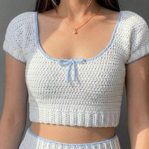 May include: A white crochet crop top with light blue trim and a small bow at the neckline. The top has short sleeves and a square neckline. The matching skirt is also visible. This handmade top is perfect for summer.