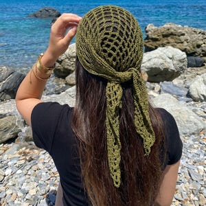 May include: A green crocheted head covering with a net-like design, tied at the back. The head covering is olive green and is worn by a person with long brown hair. The background features a rocky beach and the ocean.