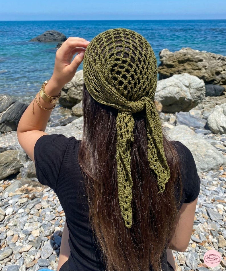 May include: A green crocheted head covering with a net-like design, tied at the back. The head covering is olive green and is worn by a person with long brown hair. The background features a rocky beach and the ocean.
