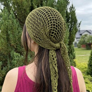 May include: Olive green crochet head covering with a net-like design. The head covering is tied at the back, with long, decorative, openwork ties. The person is wearing a pink tank top. The background is a green, leafy environment.
