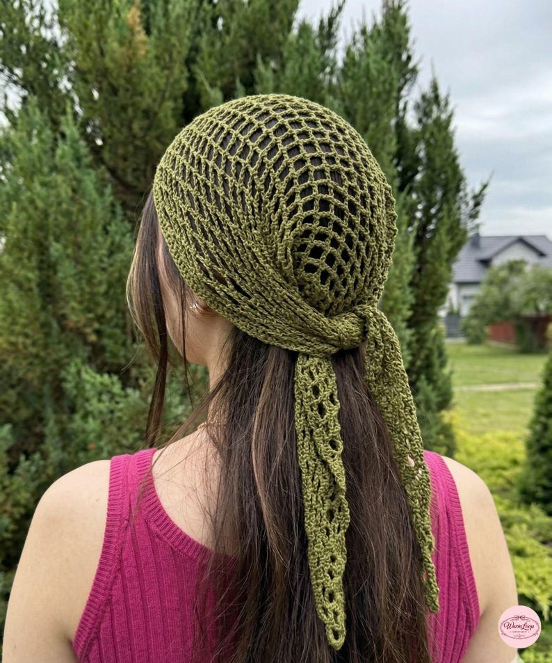 May include: Olive green crochet head covering with a net-like design. The head covering is tied at the back, with long, decorative, openwork ties. The person is wearing a pink tank top. The background is a green, leafy environment.