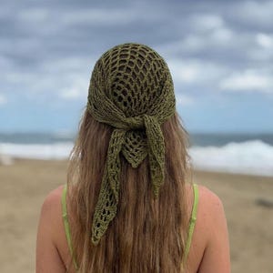 May include: Olive green crochet headscarf with a net-like design, tied at the back. The head covering is worn over long, light brown hair. The background shows a beach and ocean under a cloudy sky.
