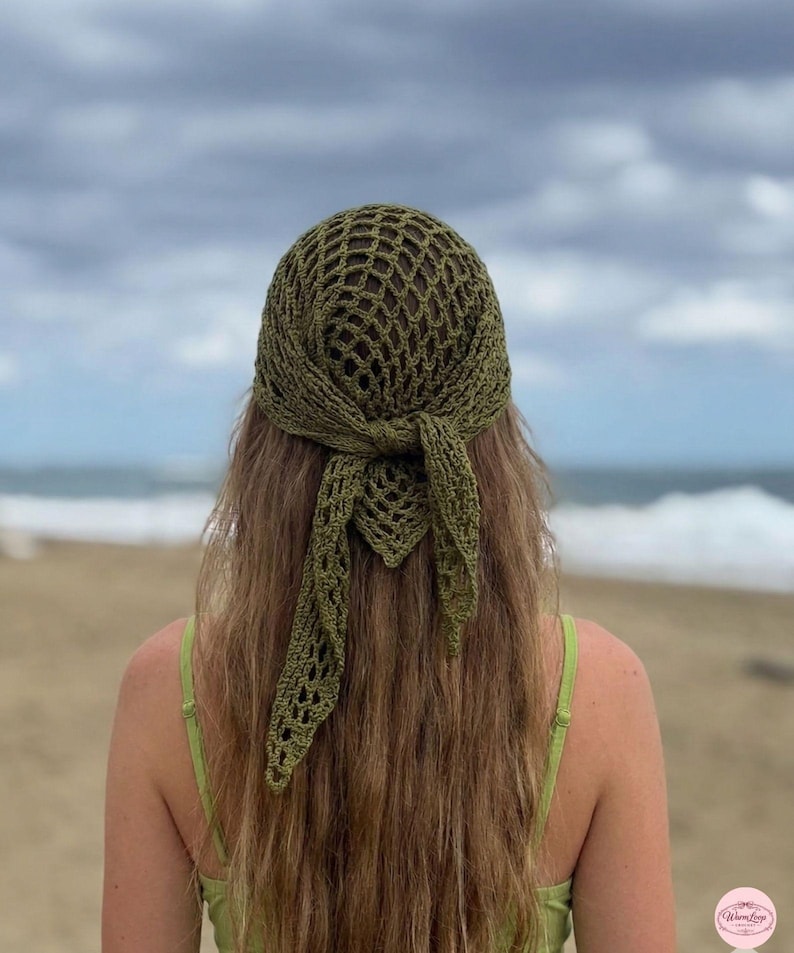 May include: Olive green crochet headscarf with a net-like design, tied at the back. The head covering is worn over long, light brown hair. The background shows a beach and ocean under a cloudy sky.