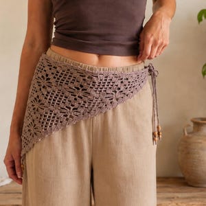 May include: A mauve crochet hip scarf featuring an intricate openwork floral pattern with a triangular shape. The garment is secured at the side with thin braided ties finished with small wooden beads, worn over light tan wide-leg trousers.