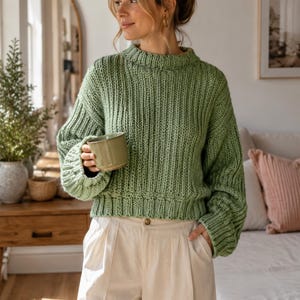 May include: A sage green chunky knit sweater with a ribbed texture, a high neckline, and long sleeves, paired with cream-colored trousers featuring a front button closure and pleats. The sweater has a cropped fit and a thick, visible knit pattern.