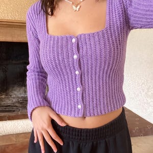 May include: A long-sleeved, lavender-colored crochet cardigan featuring a square neckline and five white, round buttons down the front. The garment has a ribbed texture throughout and is paired with black pants and a pearl necklace with a butterfly charm.