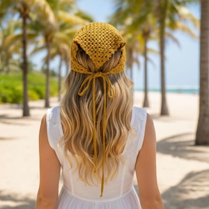 May include: Mustard yellow crochet headscarf with long ties, worn by a person with blonde hair. The headscarf is tied at the back of the head. The person is wearing a white dress. Palm trees and a beach are in the background.