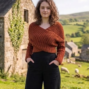 Könnte beinhalten: Eine Frau in einem rostfarbenen Wickelpullover mit langen Ärmeln und einer schwarzen Hose mit weitem Bein. Im Hintergrund befinden sich ein Steingebäude und eine grüne Wiese.