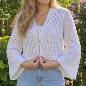 Op de afbeelding: Een witte gehaakte cardigan met uitlopende mouwen en een geschulpte rand. Het open ontwerp heeft een striksluiting bij de halslijn. De open weefconstructie maakt het een veelzijdig kledingstuk.