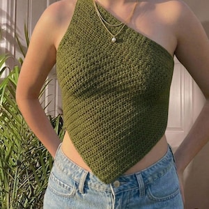 May include: Olive green crochet halter top with a triangular shape. The top features a single shoulder strap and a textured stitch pattern. A gold necklace with a pearl pendant is visible. The top is paired with blue denim jeans.