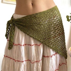 May include: A green crocheted wrap tied at the waist, draped over a cream-colored skirt with red trim. The wrap has an open, net-like design. The skirt is layered and flowing.